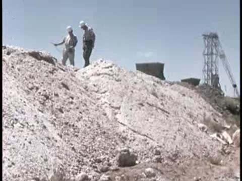 two men in hard hats standing on a rocky hill with equipment outdoors