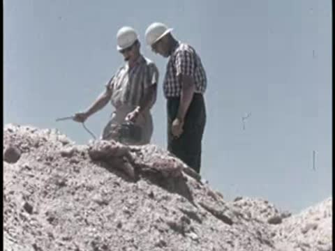 two men in hard hats standing on a rocky dirt mound outdoors