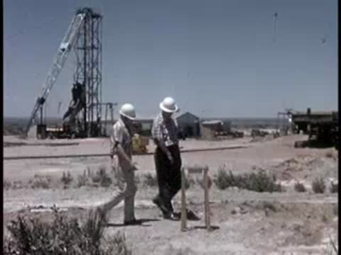 two men in hard hats walking near survey markers and a drilling rig