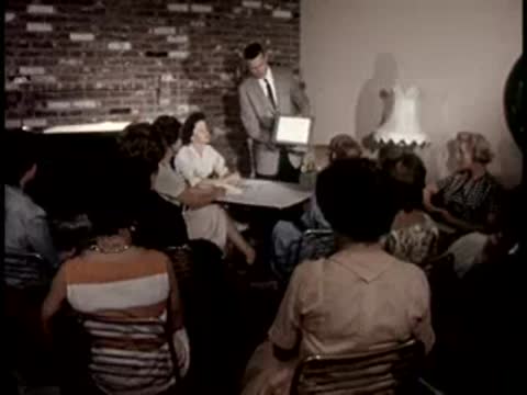 A man shows a small device to women seated in a living room, retro interior