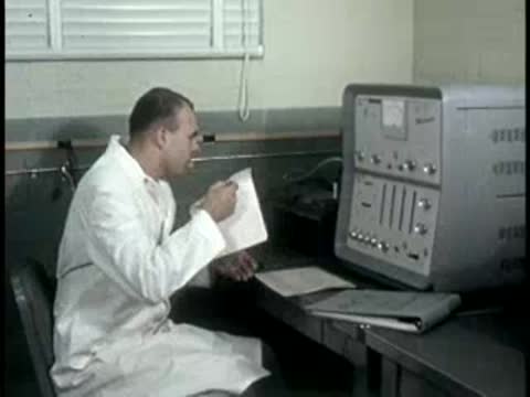 A technician in a lab coat sits at a desk, reviewing paper charts near equipment.