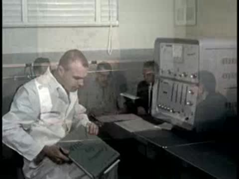 A technician at a desk interacts with equipment and a group meets at a table.