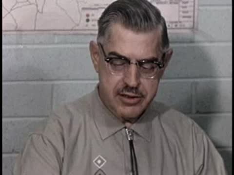 A man with glasses and a patterned shirt speaks in front of a brick wall.
