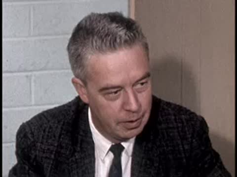 A man in a suit jacket looks down and speaks during an interview.