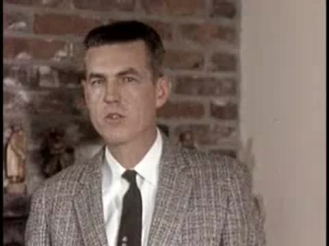 A man speaks in front of a brick wall, mid-century professional setting