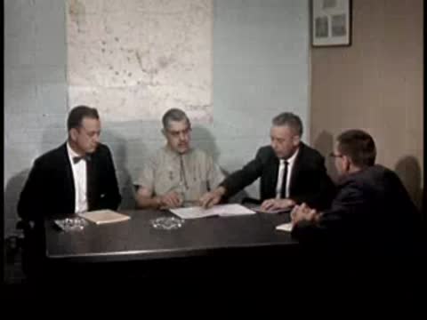 Four men in suits and uniforms sit around a conference table discussing papers.