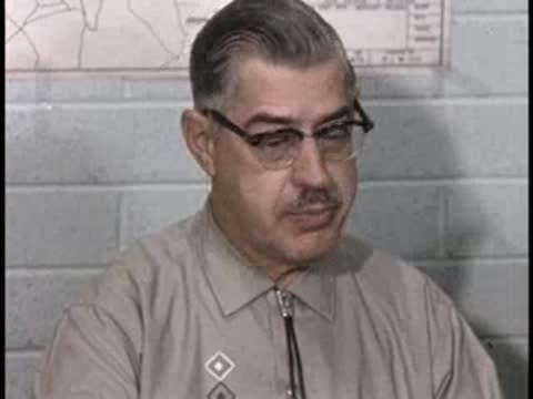 A man with glasses and a patterned shirt speaks animatedly in front of a wall.