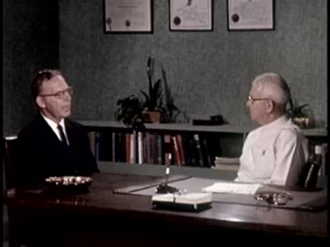 A man in a dark suit talks with a man in a white coat at a desk.