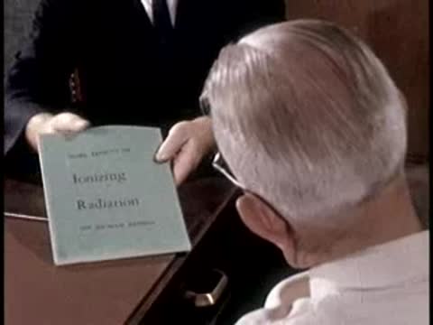 A man in a suit hands a light blue book titled "Ionizing Radiation" to a doctor.