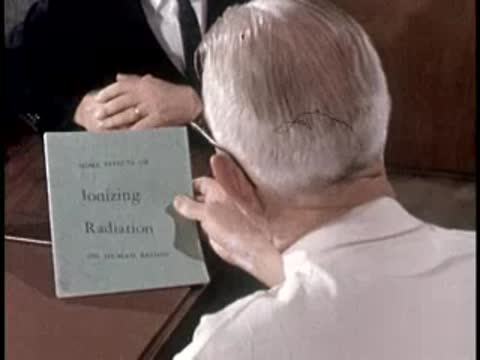 A doctor in a white coat opens a blue book titled "Ionizing Radiation" at a desk.