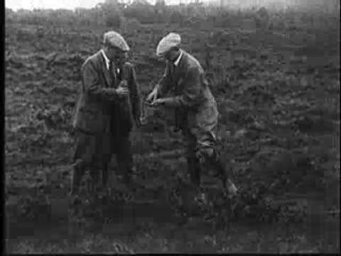 Three men in vintage clothing stand in a barren field, one leaning down.
