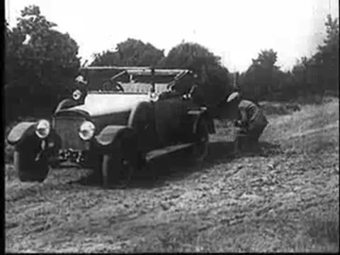 Vintage car stopped on dirt road; men unloading camouflage gear from the back.