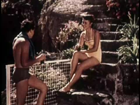 A man and woman relaxing on stone steps near the water, mid-century swimwear.