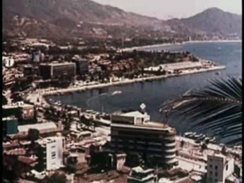 An aerial view of a coastal city with hills and buildings, mid-century.