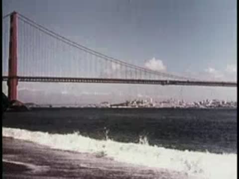 Waves crashing on a beach near a bridge and city, 1950s.