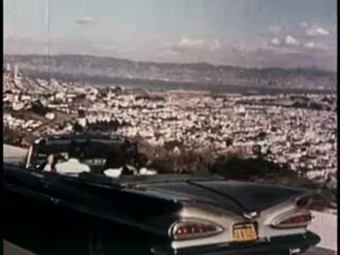 A convertible car driving along a scenic road overlooking a city, 1950s.