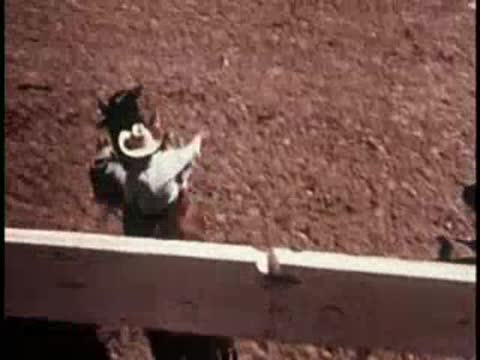 A rider on a horse jumping out of a gate into a dirt arena.