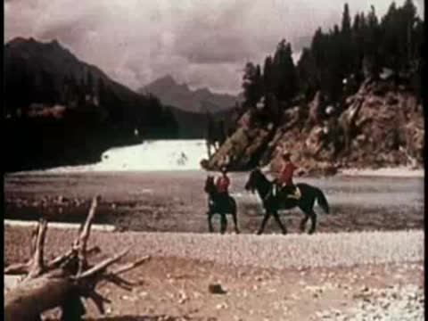 two people on horseback near a wide river with forested mountains in the background