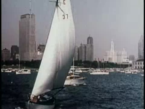 sailboat with a large white sail on a busy harbor with city skyscrapers behind