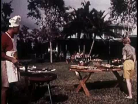 people interacting around an outdoor barbecue area with tables and tropical trees